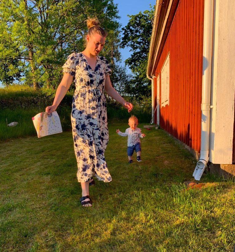 Kvinna i blommig klänning går på gräs med ett barn som springer framåt.