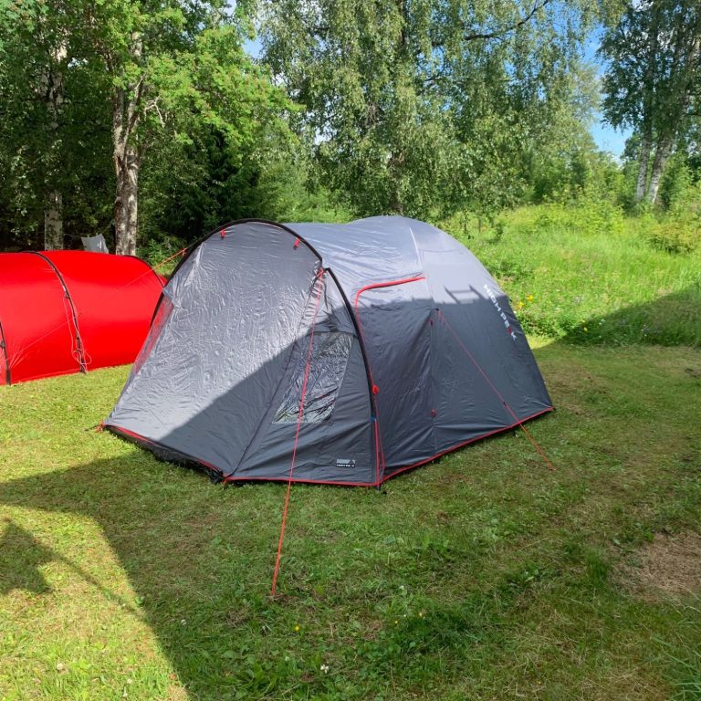 Grått tält med röda detaljer på grön gräsyta omgiven av träd.