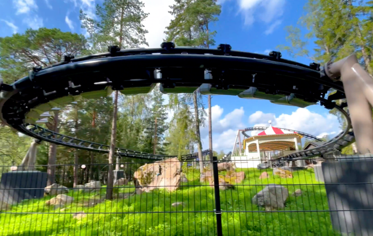 Svart rutschkana i ett grönskande område med träd och en byggnad i bakgrunden.