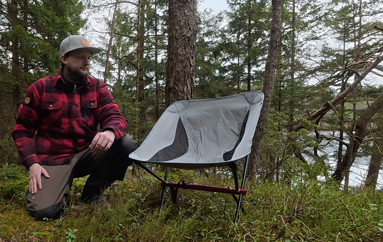 Man in a forest next to a portable camping chair by a lake.
