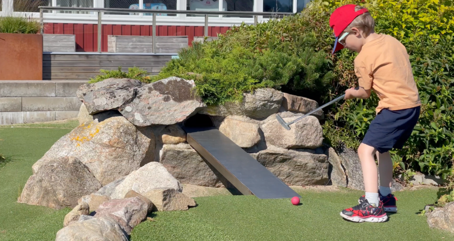 Barn spelar minigolf med en klubba, placerad bakom stenar och en ramp.
