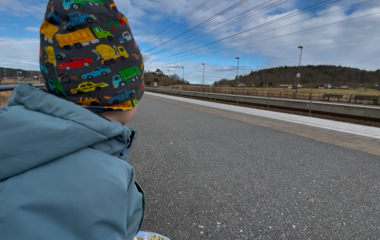 Barn i färgglad mössa sitter på en plattform med tågspår i bakgrunden.