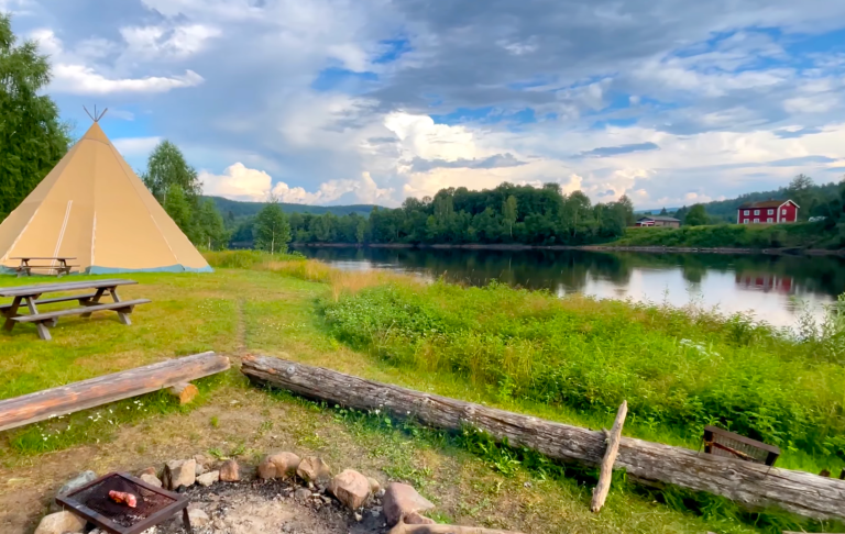 Lägerplats vid en sjö med ett tält, bänkar och en lägereld under en molnig himmel.
