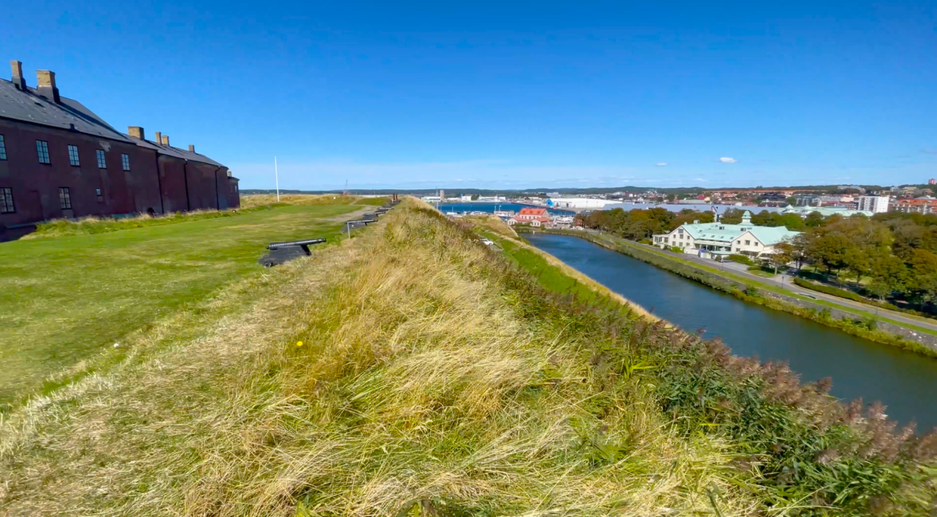 Varbergs Fästning Gröna åkerkanter och en flod med båtar, omgiven av byggnader och himmel.