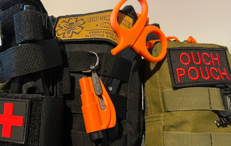 Svart och grön medicinväska med orange sax och nyckelring. Texten "OUCH POUCH" synlig.