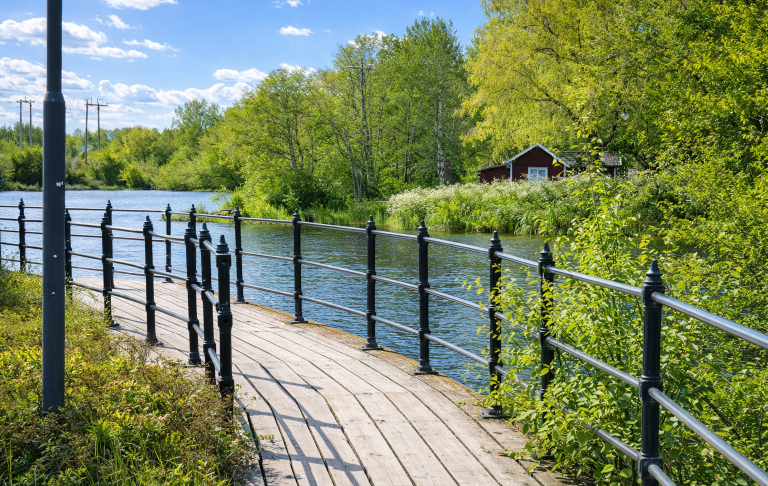 En träpromenad längs med en lugn flod, omgiven av grönska och träd.
