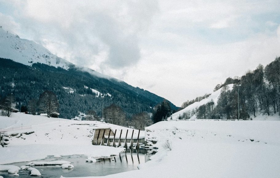 Snöiga berg med en frusen sjö och en träbro i förgrunden.