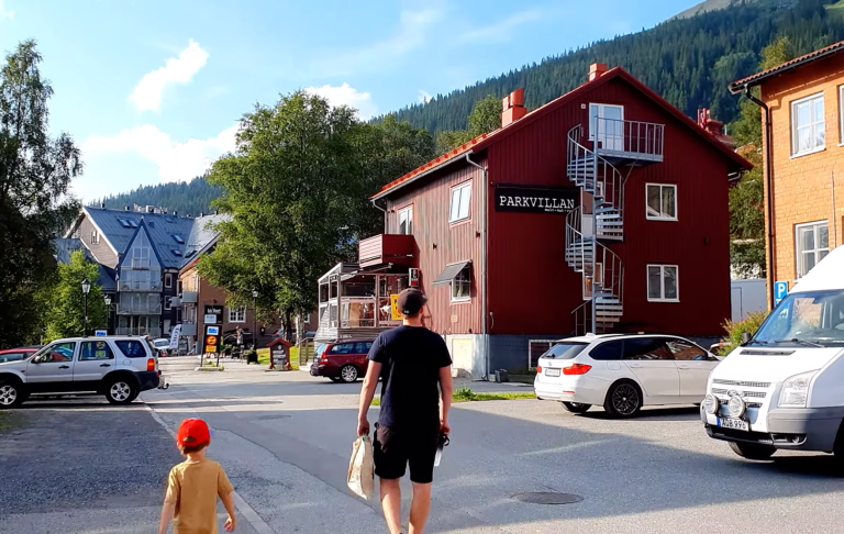 Två personer går på en gata med röd träbyggnad och bilar parkerade.
