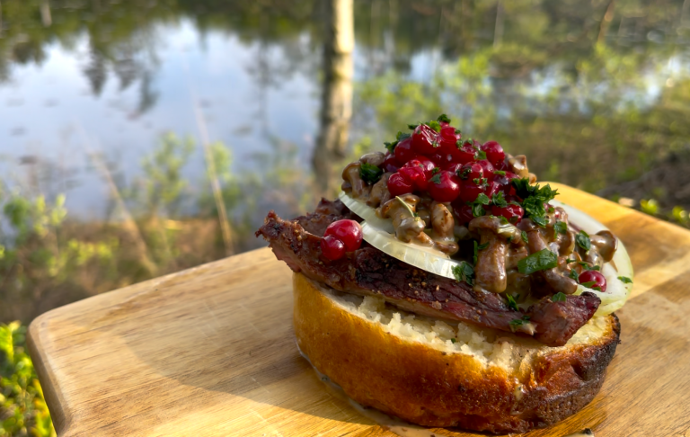 Vildmarkssmörgås med högrev och kantareller En saftig smörgås med kött, lök, kantareller, lingon och örter, utomhus vid en sjö.