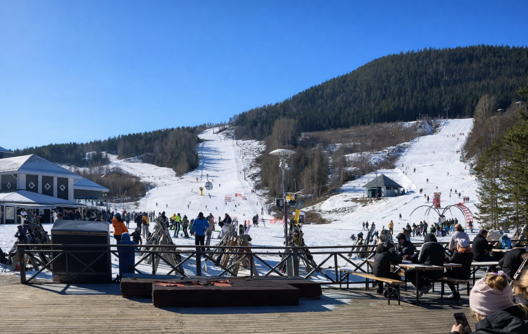 Skidbacke med snö, människor åker skidor och snowboard under en klarblå himmel.