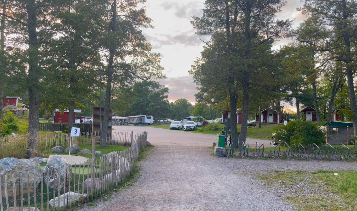 Trosa Havsbad & Camping Grusväg genom en campingplats med träd, stugor och bilar i bakgrunden.
