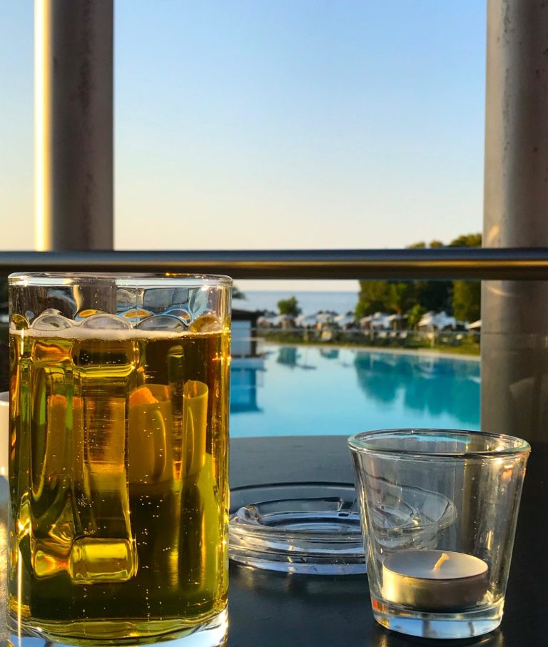 Glas med öl och ett litet glas vid en pool med havsutsikt i solnedgång.