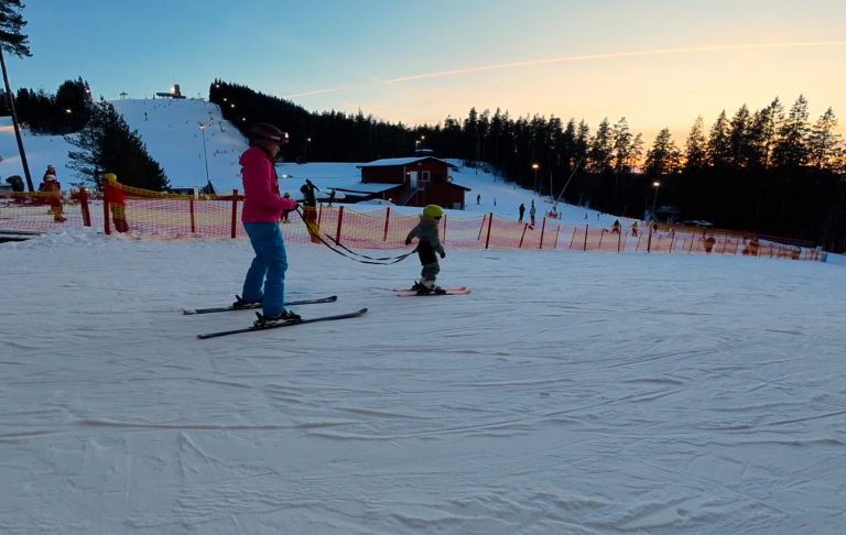 En vuxen och ett barn åker skidor på en backe vid solnedgången.