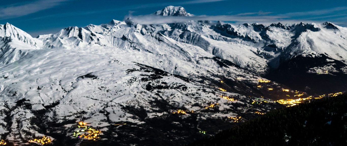 Snötäckta berg med en fjärran topp och en belyst dal i skymningen.