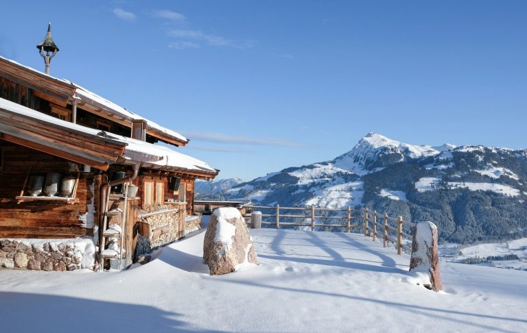 Stuga i snöigt landskap med berg i bakgrunden och klarblå himmel.