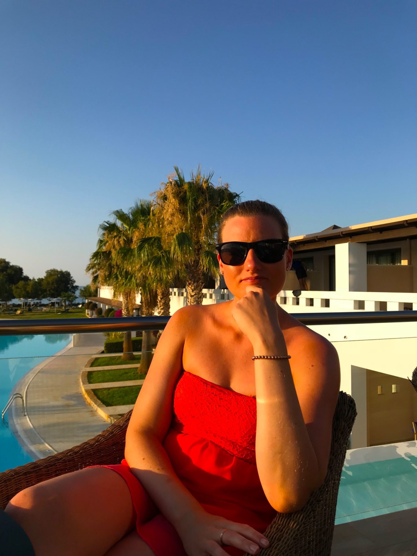 Rosita på Middag Kvinna i röd klänning och solglasögon sitter vid poolen under klar blå himmel.