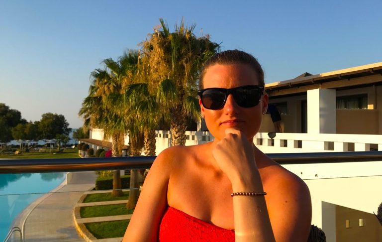 Kvinna med solglasögon vid en pool, iförd röd klänning och kort hår.