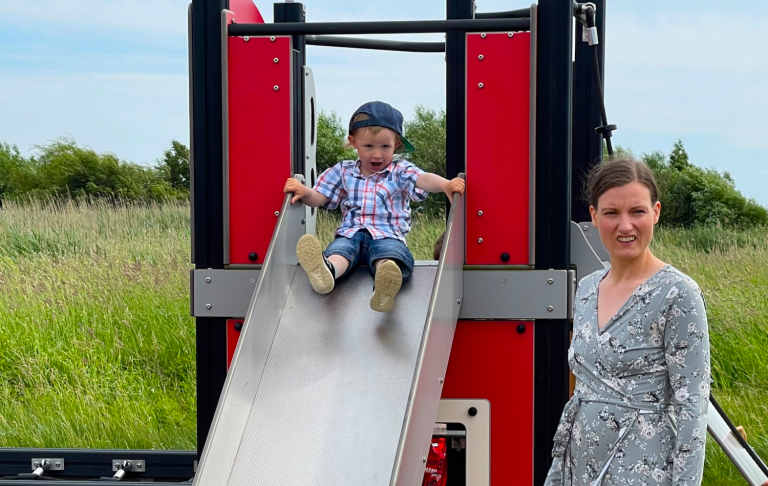 Barn åker ner för en rutschkana medan en vuxen står bredvid. Grönskande landskap i bakgrunden.