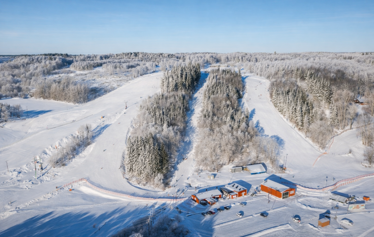 Snötäckt skidbacke med stugor i en vinterlandskap. Granskog i bakgrunden.
