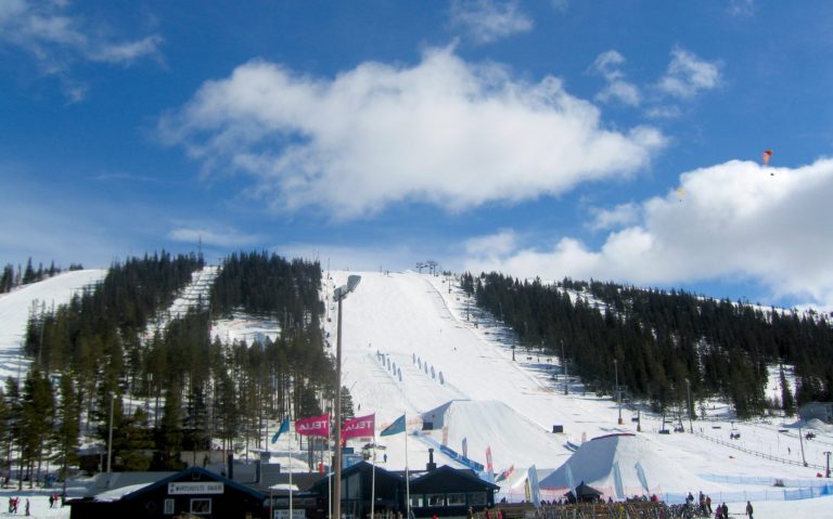 Snöklädda skidbackar med träd och blå himmel, flaggor syns i förgrunden.