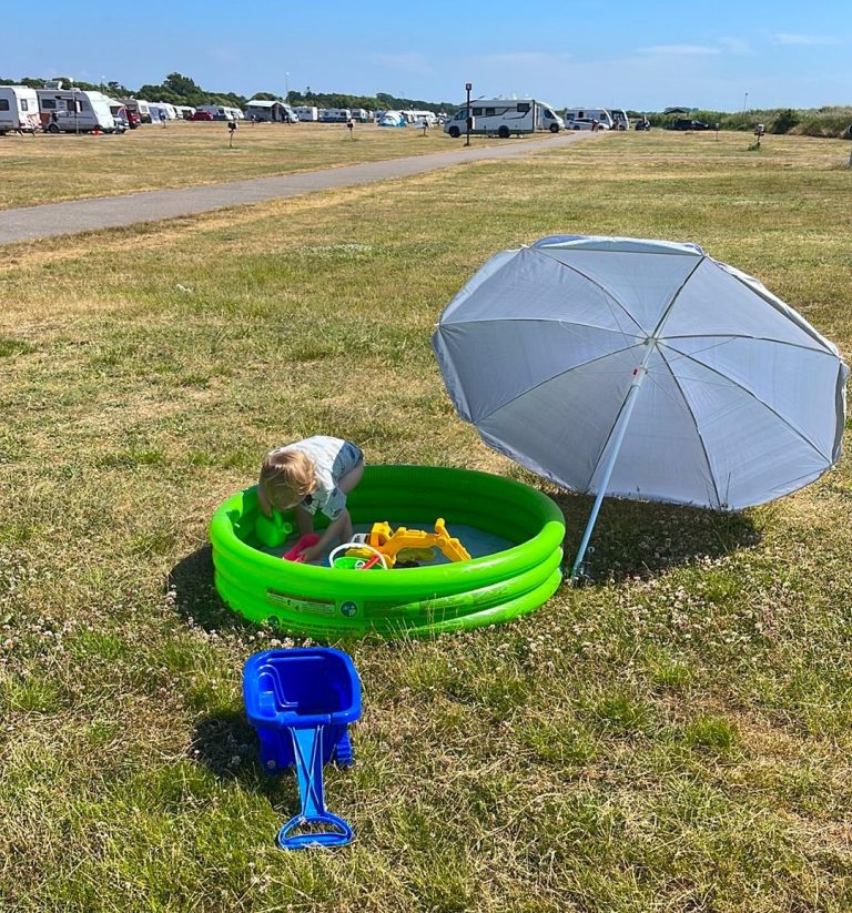 Ett barn leker i en grön plastpool under ett parasoll på en campingplats.
