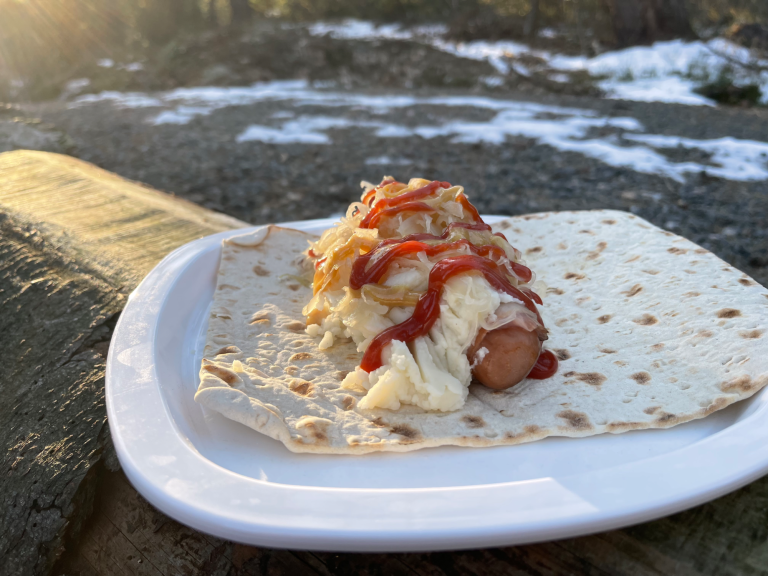 Varmkorv med potatismos, senap och ketchup på ett tunnbröd. ute i vildmarken, snö i bakgrunden.