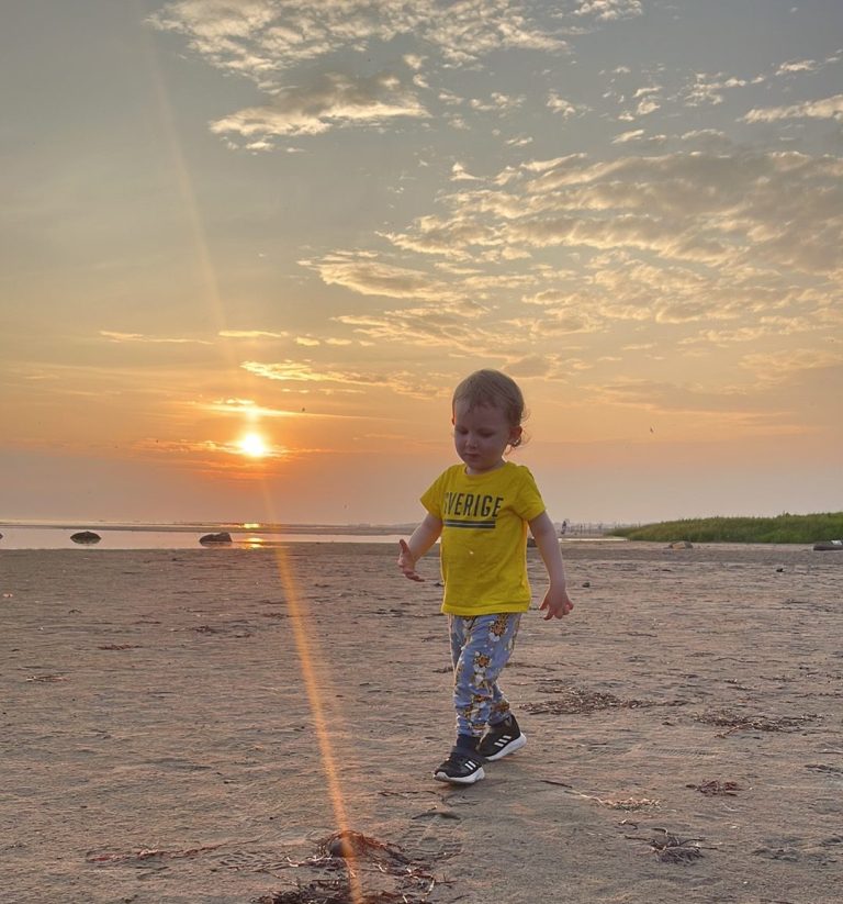 Ett barn i gul t-shirt går längs en strand vid solnedgång.