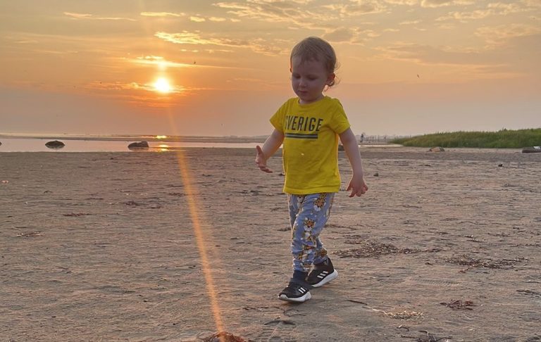 Litet barn går på strand vid solnedgång, iförd gul tröja och byxor med motiv.