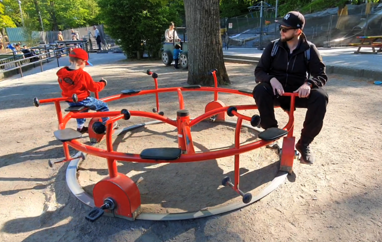 Leker på plikta i slottsskogen Två personer vid en snurrande lekutrustning i en park. Soligt väder.