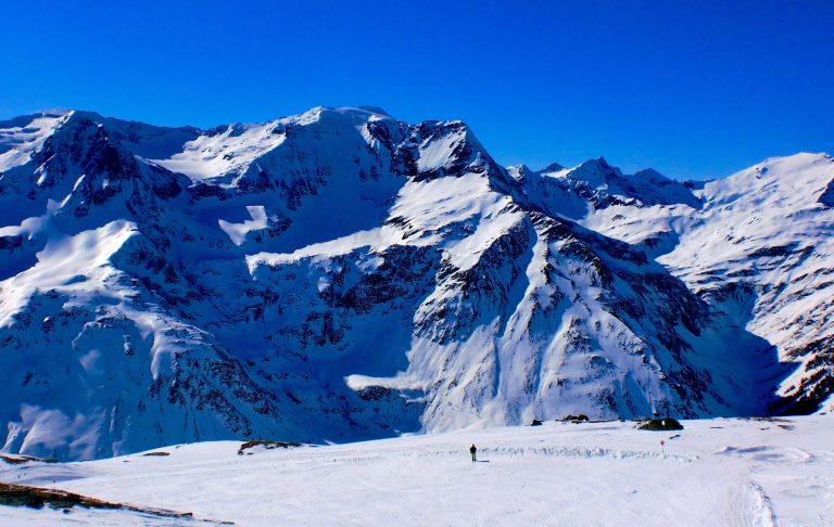 Snöklädd bergskedja med granar och en frusen sjö i förgrunden. Klara blå himmel.