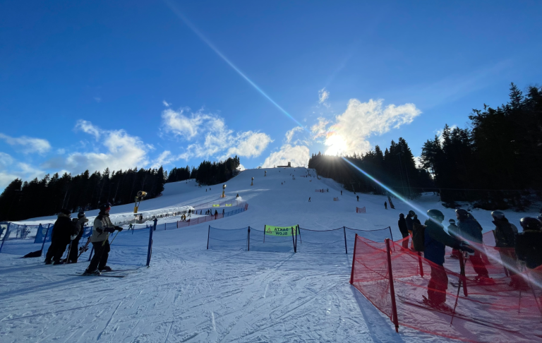 Snöig skidbacke med träd och människor i vintersportkläder. Solen strålar bakom berget.