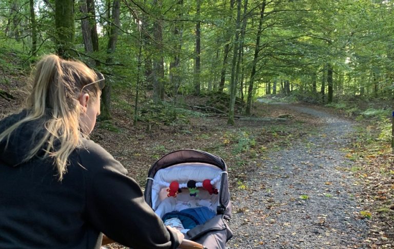 Kvinna med barnvagn på grusig stig i en skog. Soligt och grönt omkring.