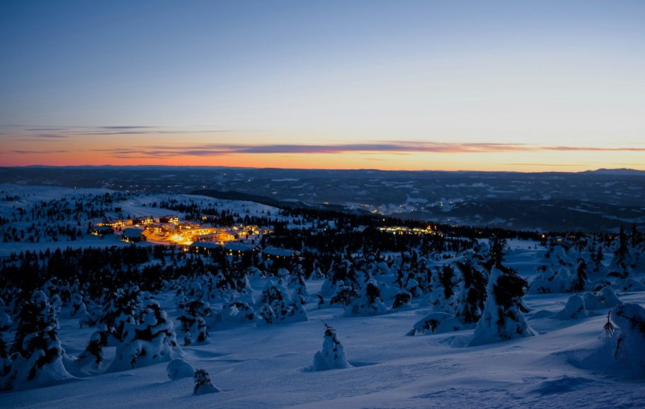 Vinterlandskap med snöklädda träd och en solnedgång över en glödande by.