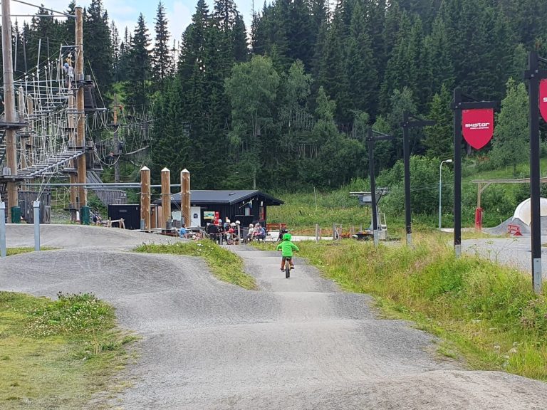 Cyklist i grön tröja på grusväg, omgiven av träd och byggnader.