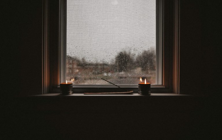 Två ljus tända på en fönsterbräda med regnig utsikt utanför.