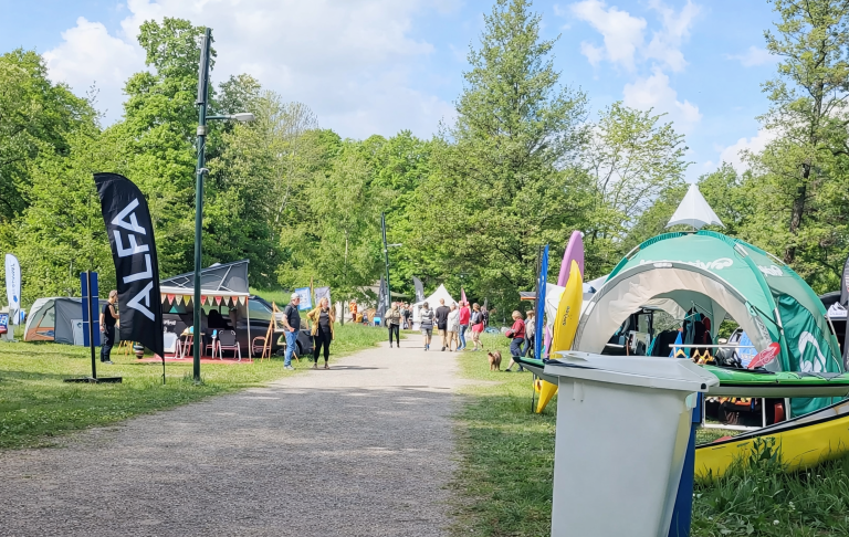 En Outdoorfestival Festivalområde med tält, bås och besökare i parkmiljö. Grönska och blå himmel.
