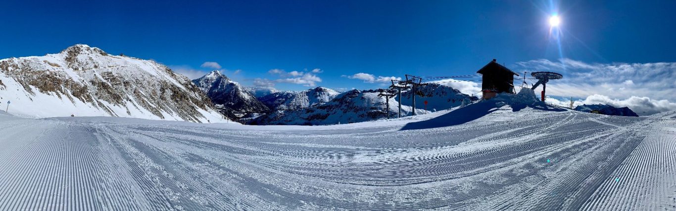 Vinterlandskap med snötäckta alpberg, skidbacke och en liftstation under en klar blå himmel.