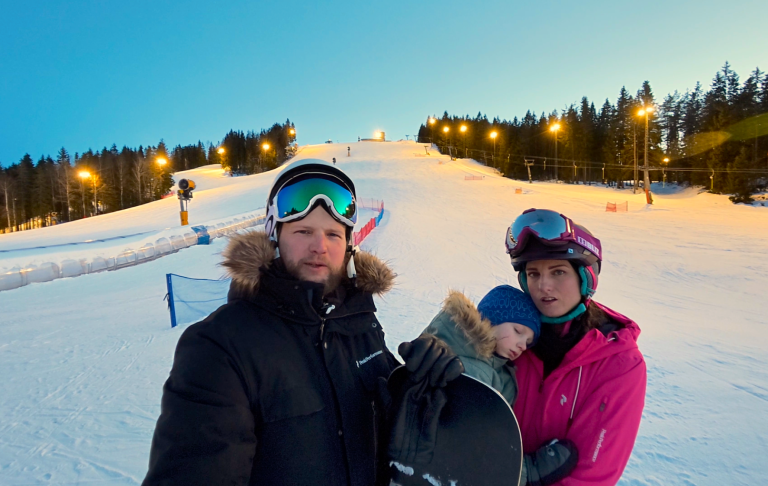 En man och en kvinna med skidutrustning på en snötäckt backe vid kvällsljus.