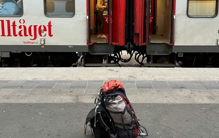 Resenär med ryggsäck väntar vid tågstation framför ett tåg som står stilla.