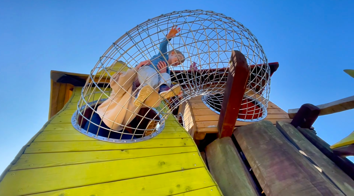 Vallarnas Friluftsområde Lekplatsen En färgglad lekplats med en nätad klättringsstruktur och en figur i buren.