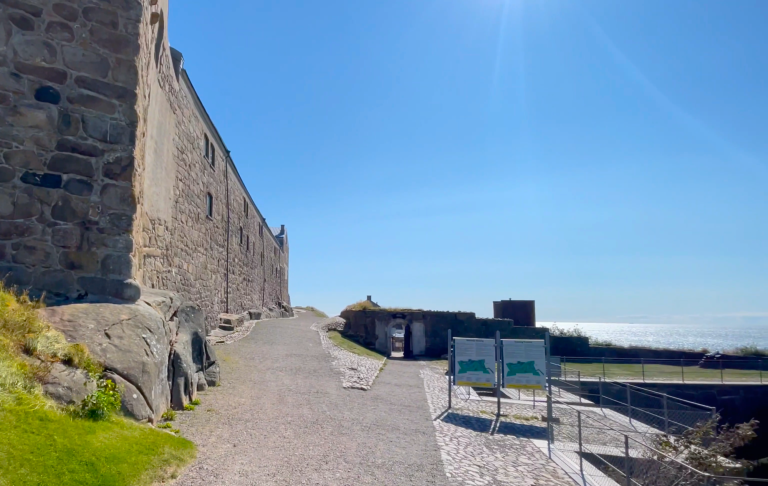 Klar blå himmel och stenmur med gångstig vid ett fort vid havet.