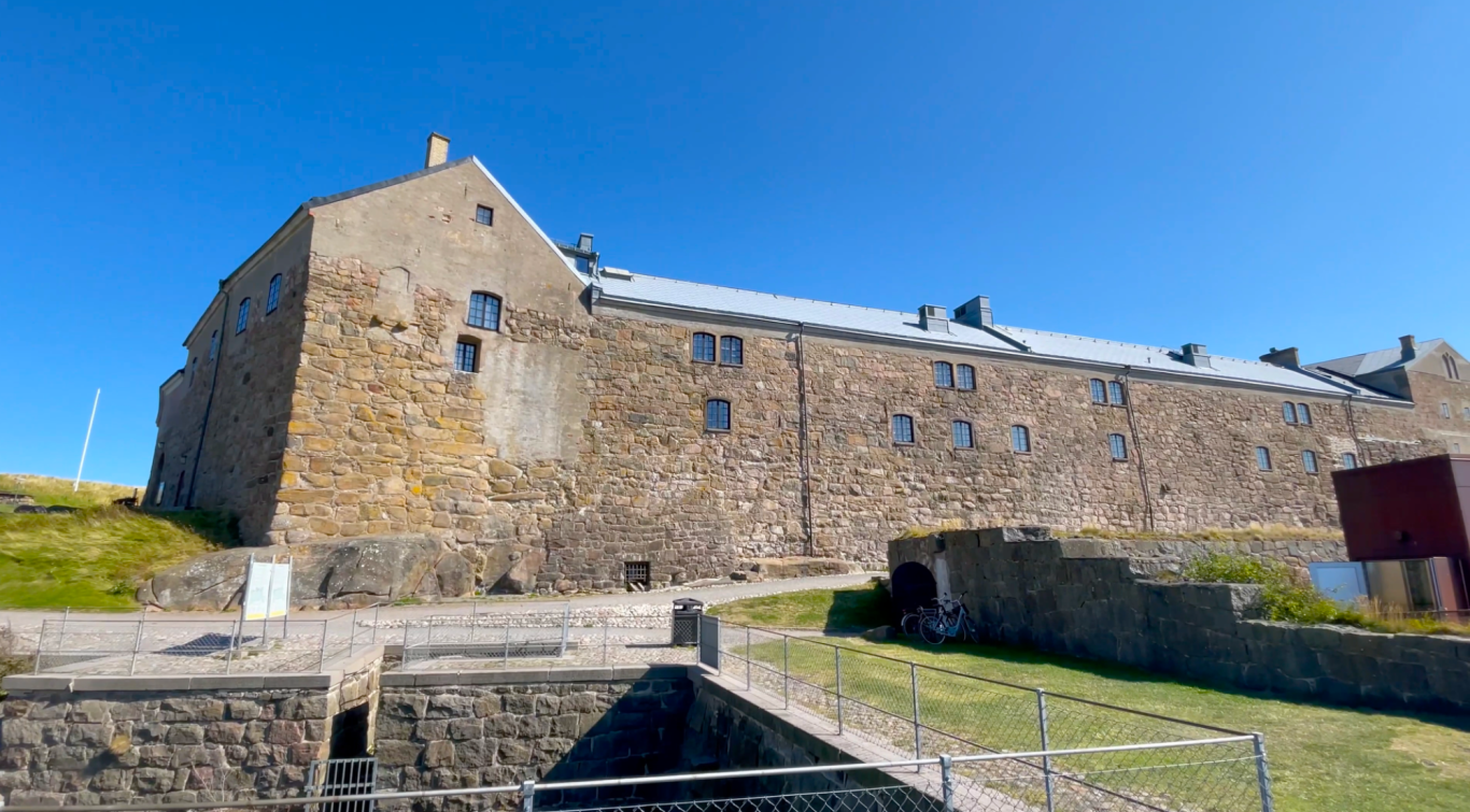 Varbergs Fästning Stenbyggnad med tak och grönsaksfält i förgrunden under klarblå himmel.