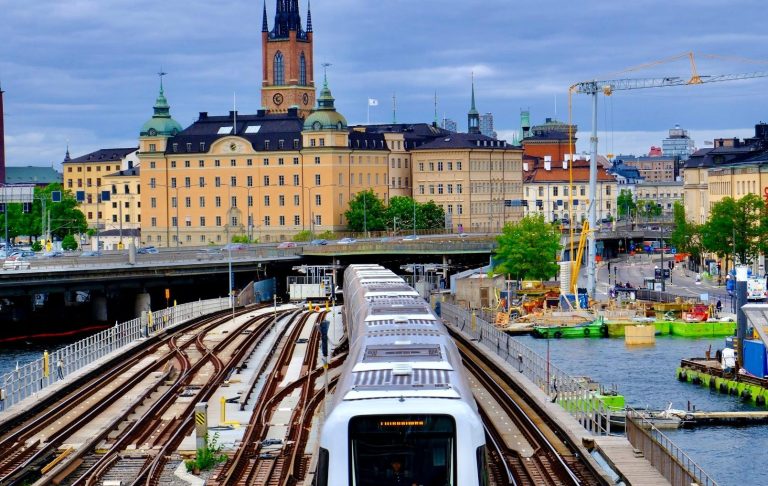 Ett tåg passerar en stad med byggnader och vatten i bakgrunden.