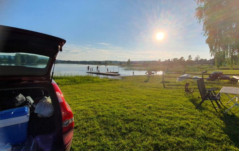Solig strand med grön gräsmatta, bilbagage öppen och två båtar på vattnet.