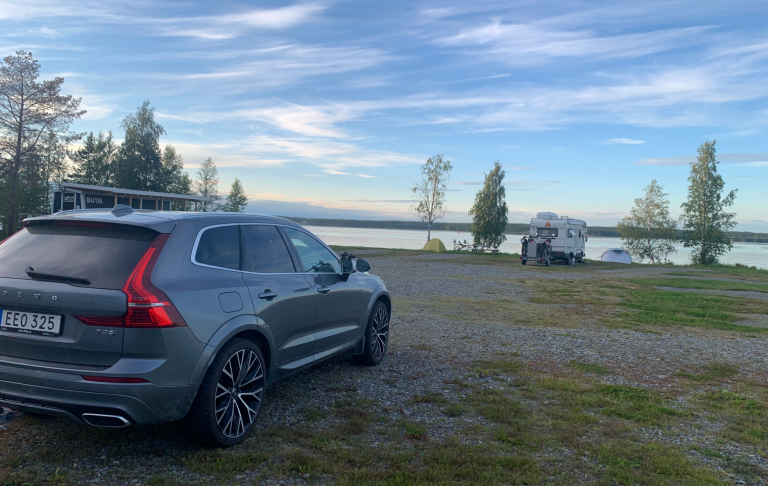 Stopp längsvägen en bilsemester Grå SUV parkerad vid en sjö med grönska i bakgrunden och en husvagn längre bort.