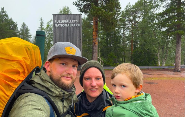 Tre personer i regnkläder med en ryggsäck i en skogsmiljö.