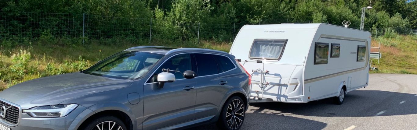 En grå bil kopplad till en vit husvagn, parkerad vid en grönskande plats.