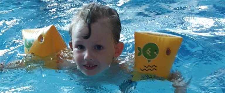 Ett barn i vatten med gula simhjälpmedel, ler och ser glad ut.
