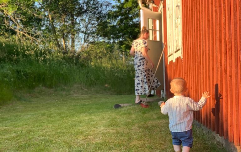 Ett barn i vit tröja går mot en vuxen som arbetar vid en röd stuga i soligt väder.