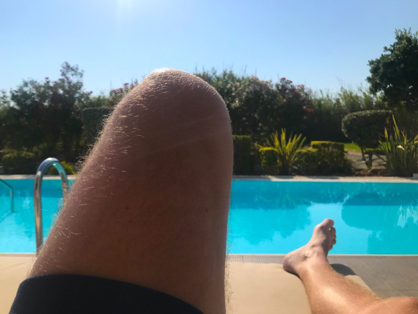 Johannes vid poolen Närbild av ett ben vid en pool med soligt väder och grönska i bakgrunden.